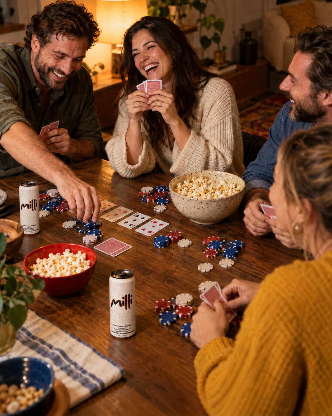 Friends laughing around a poker table with chips, popcorn and cans of Milli