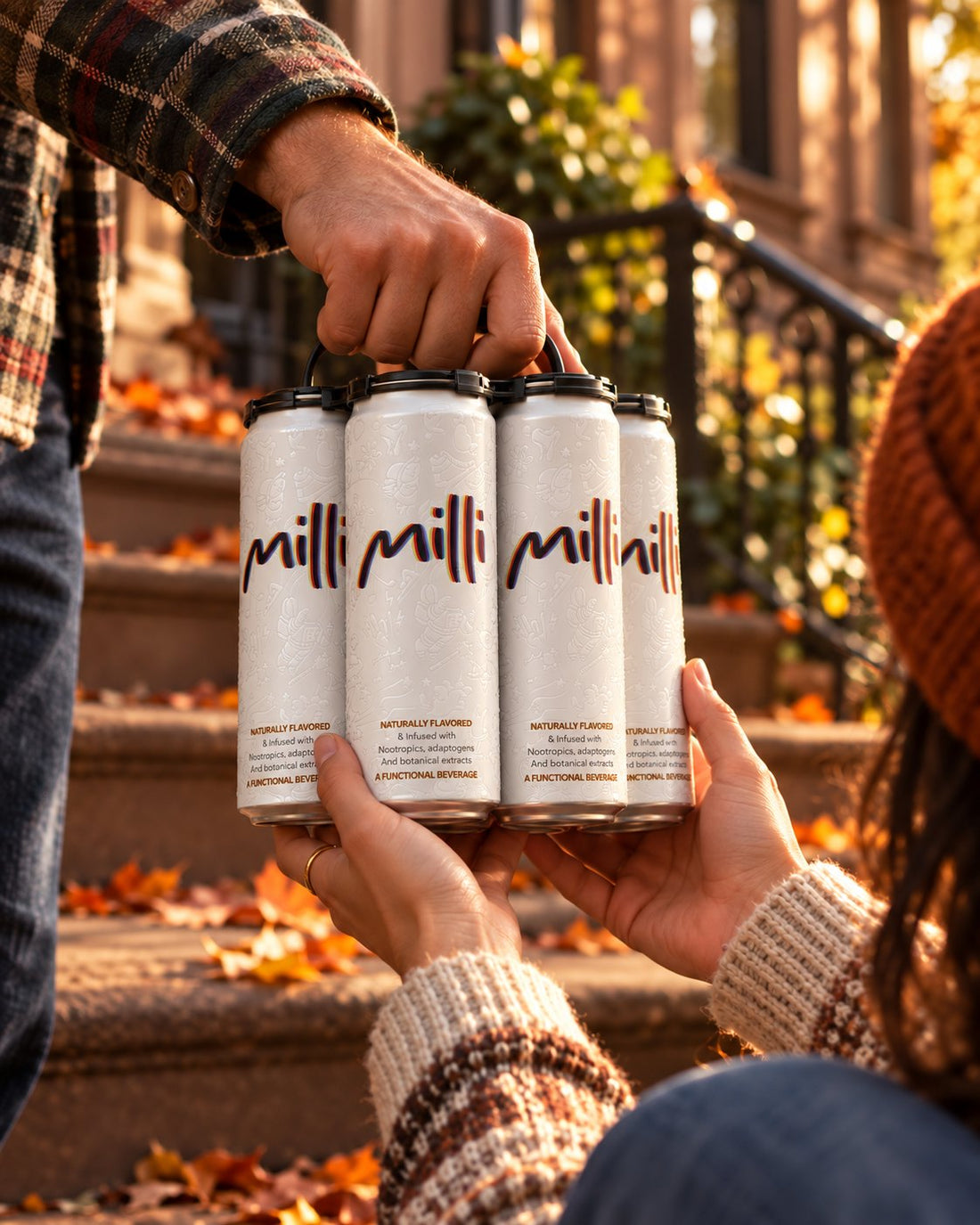 Hands pass a 4-pack of Milli cans on a stoop covered in fall leaves
