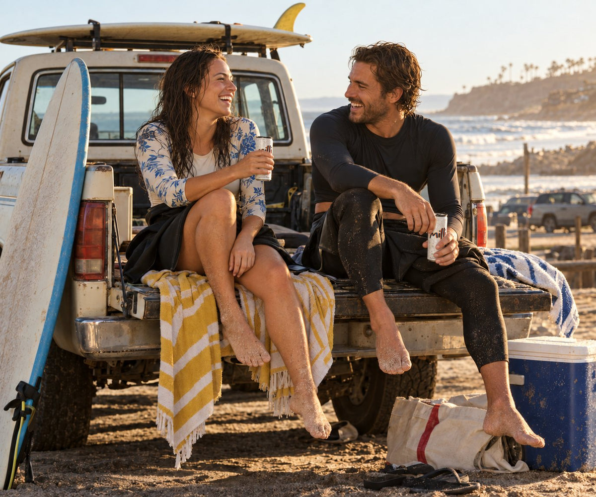 A surfer couple sits on a truck tailgate by the beach, each with a can of Milli