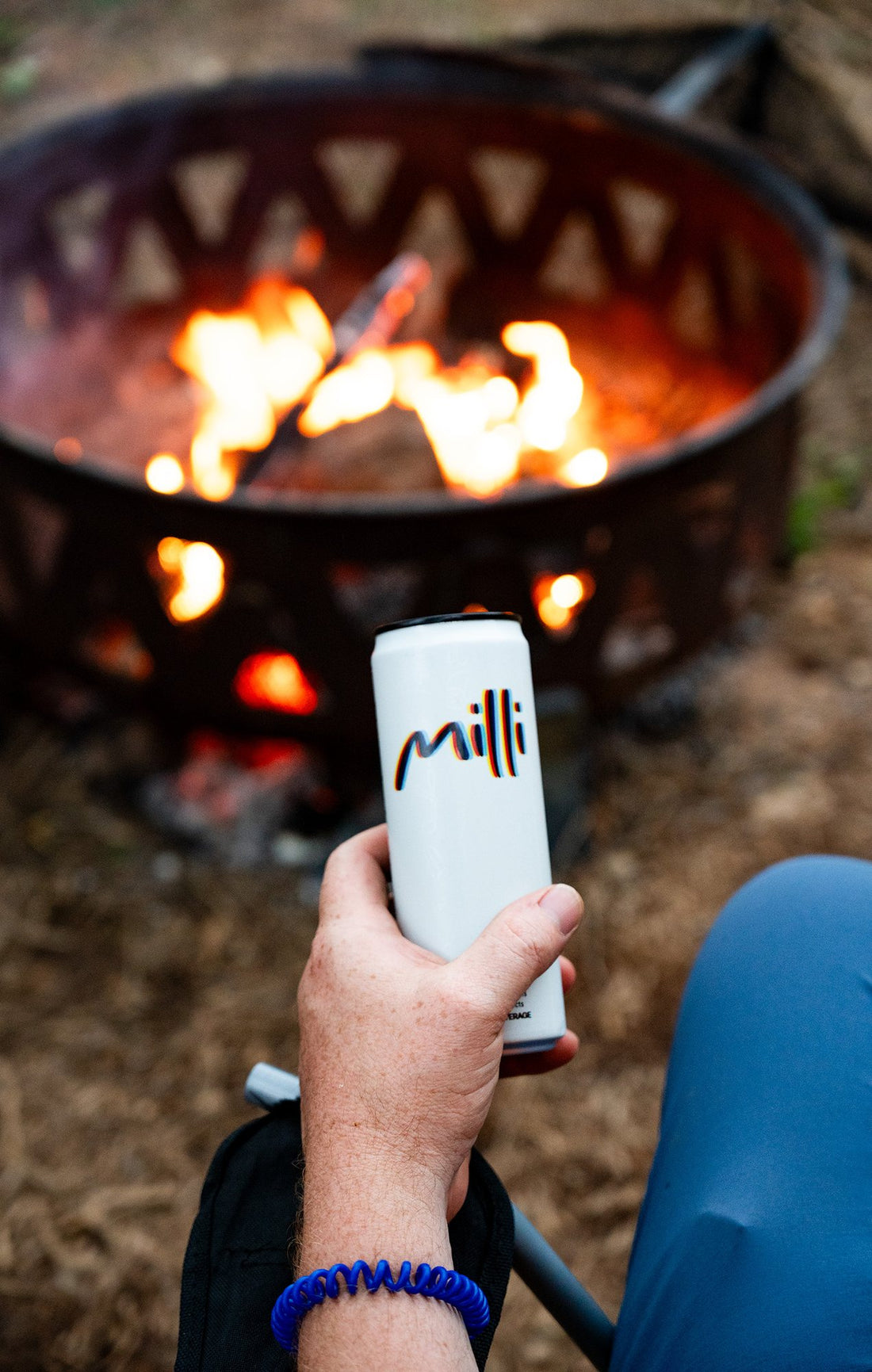 A hand holds a can of Milli beside a glowing fire pit