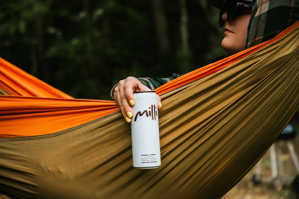 Someone relaxing in an orange hammock in the woods holds a can of Milli