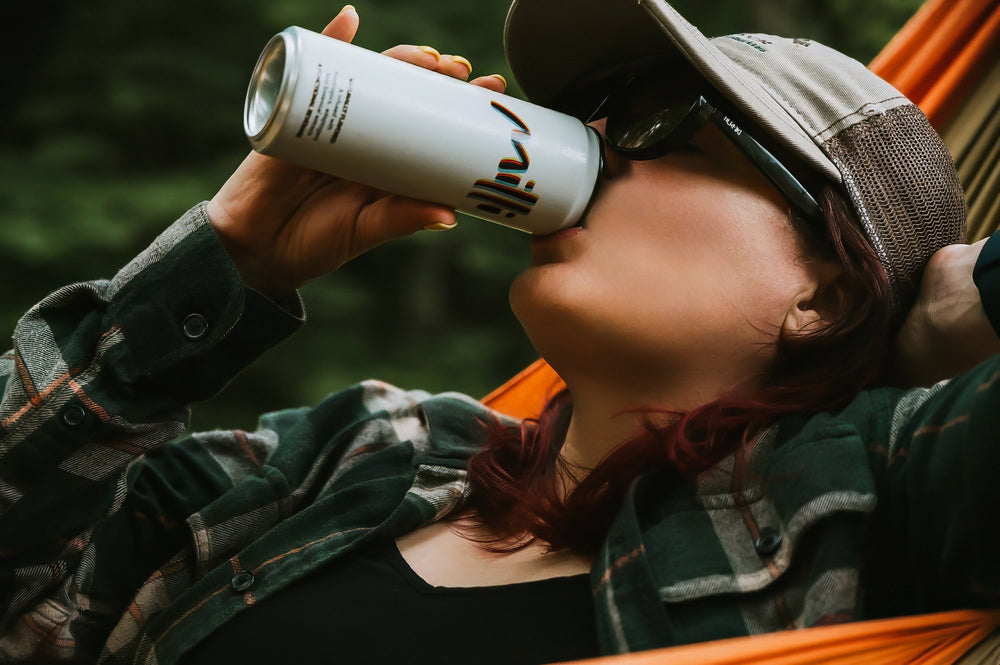 A woman in a cap and flannel shirt sips a can of Milli while lying in a hammock