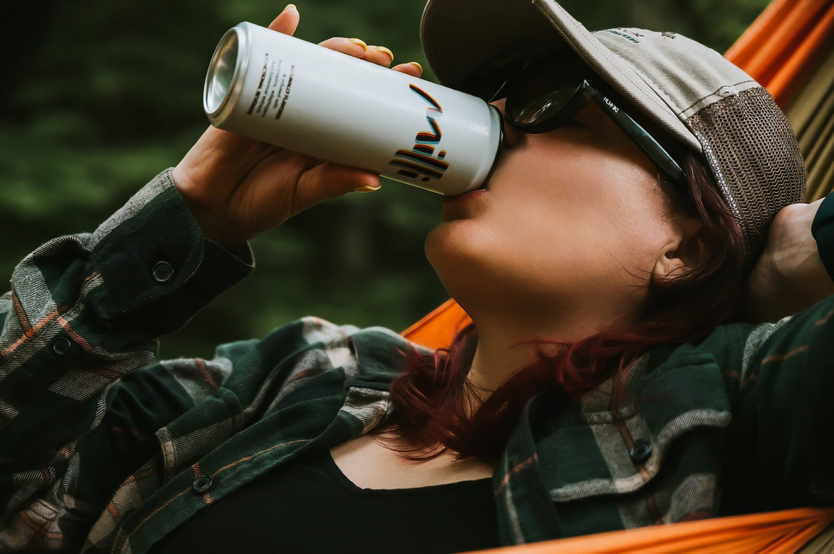 A woman in a cap and flannel shirt sips a can of Milli while lying in a hammock